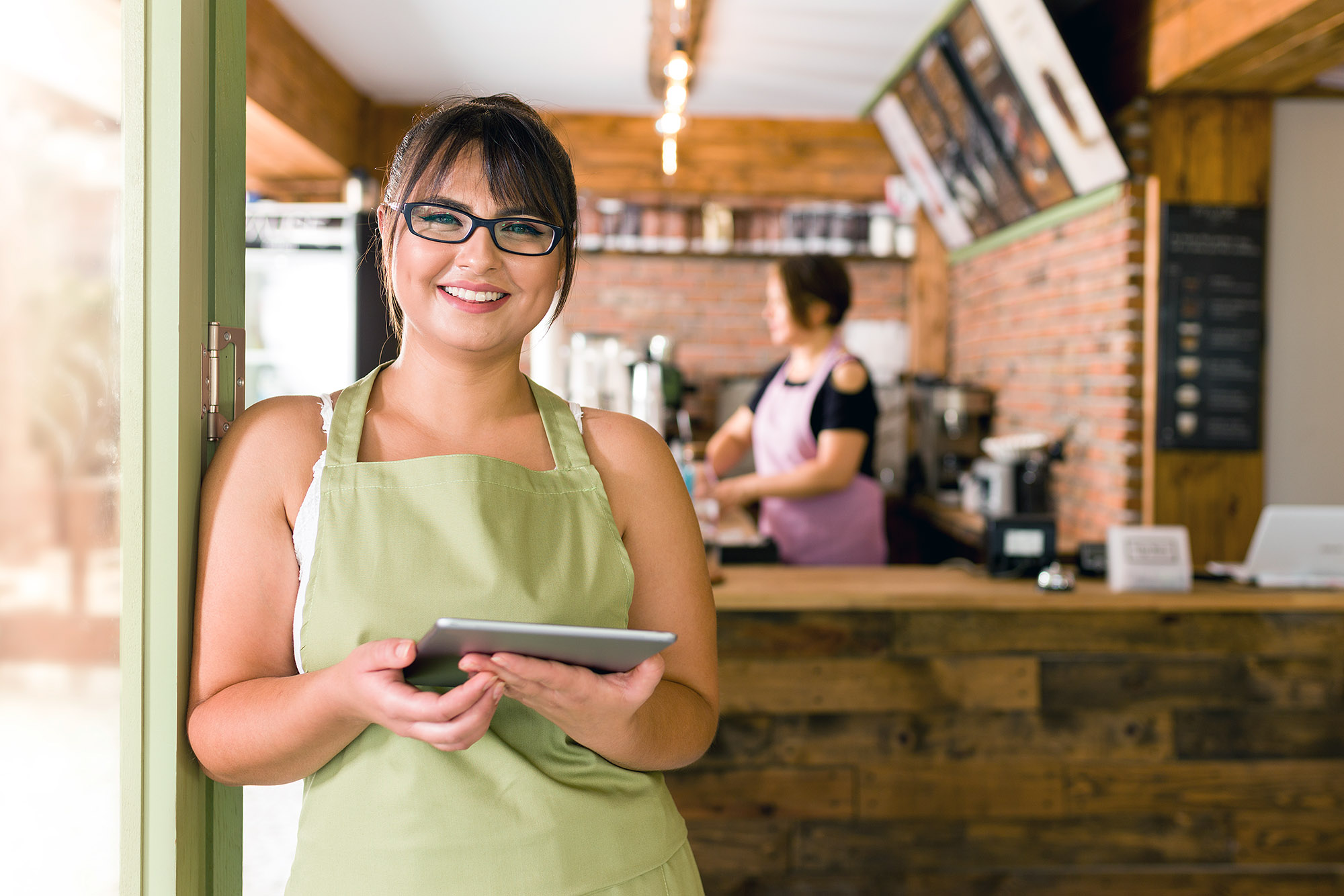 A happy restaurant owner stands at the door of her shop holding a tablet and smiles